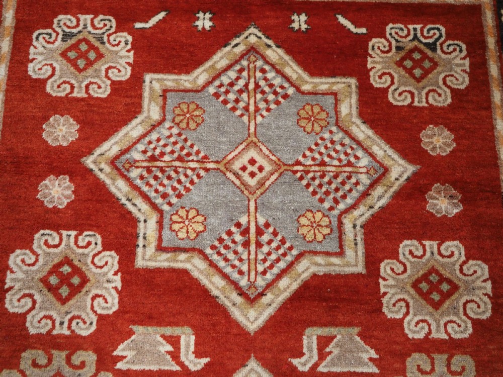 An antique East Turkestan Khotan wool area rug featuring three light blue geometric star medallions on a red field within wide cream and beige patterned borders. View VIII