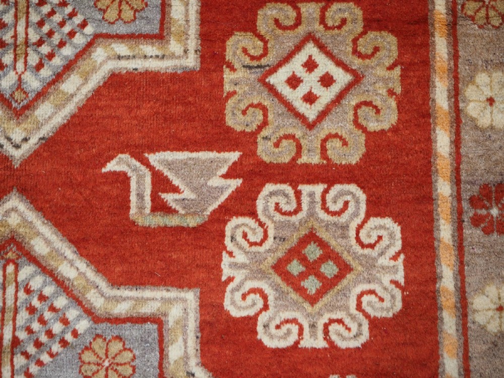 An antique East Turkestan Khotan wool area rug featuring three light blue geometric star medallions on a red field within wide cream and beige patterned borders. View IX