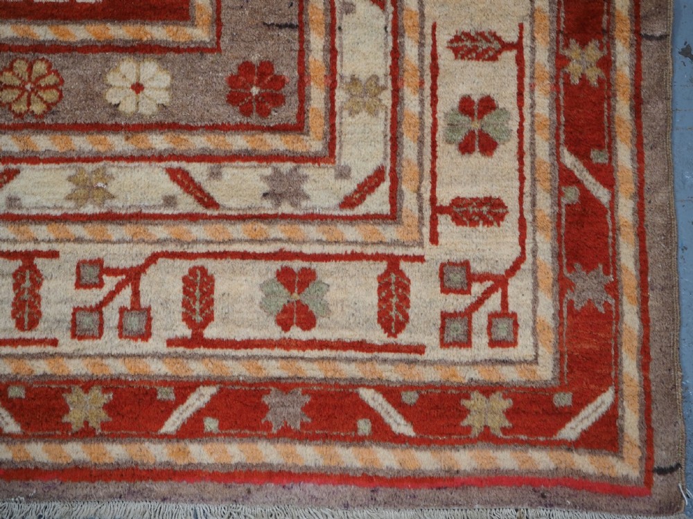 An antique East Turkestan Khotan wool area rug featuring three light blue geometric star medallions on a red field within wide cream and beige patterned borders. View XII