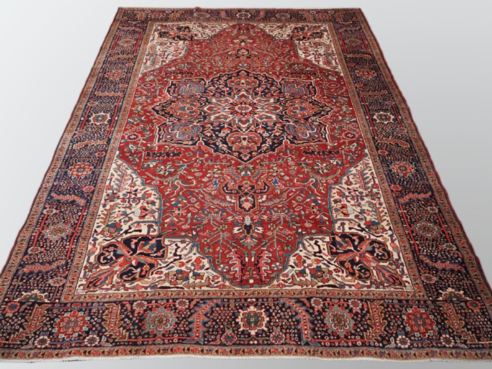 An antique hand-knotted Persian Heriz rug featuring a large geometric central medallion in navy blue, set upon a red field with off-white corner spandrels and a wide decorative border.