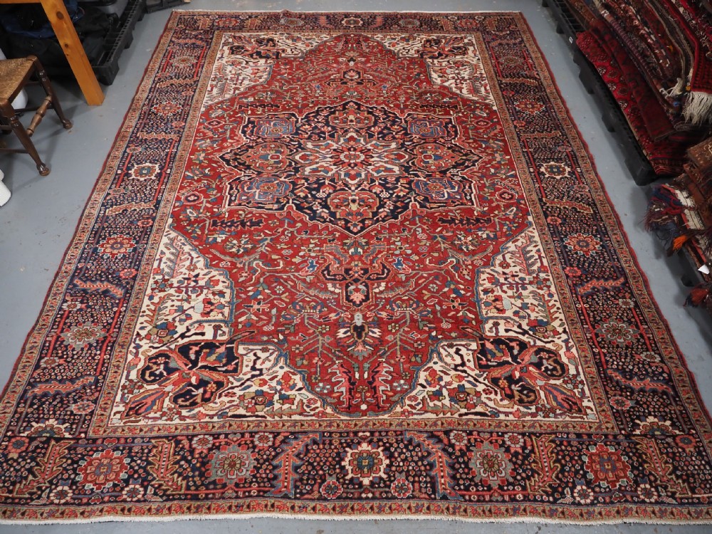 An antique hand-knotted Persian Heriz rug featuring a large geometric central medallion in navy blue, set upon a red field with off-white corner spandrels and a wide decorative border. View III