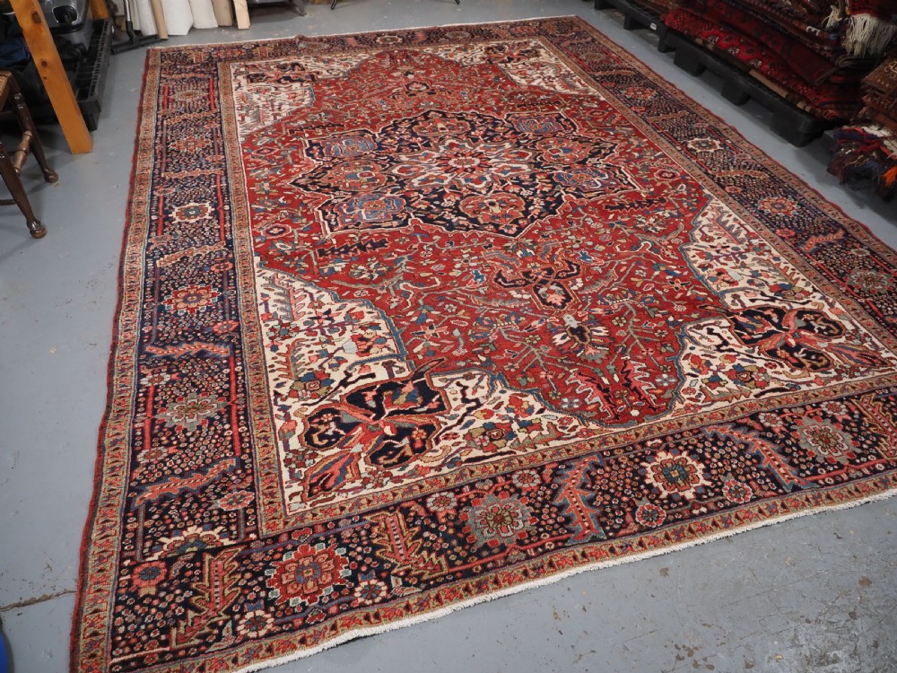 An antique hand-knotted Persian Heriz rug featuring a large geometric central medallion in navy blue, set upon a red field with off-white corner spandrels and a wide decorative border. View IV