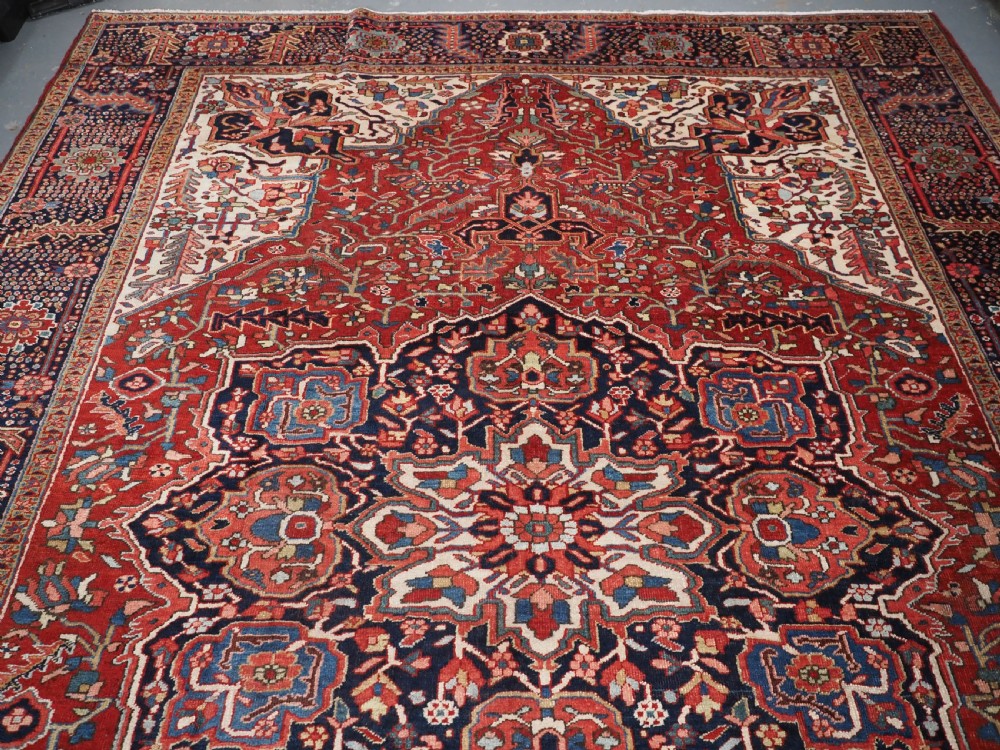 An antique hand-knotted Persian Heriz rug featuring a large geometric central medallion in navy blue, set upon a red field with off-white corner spandrels and a wide decorative border. View V