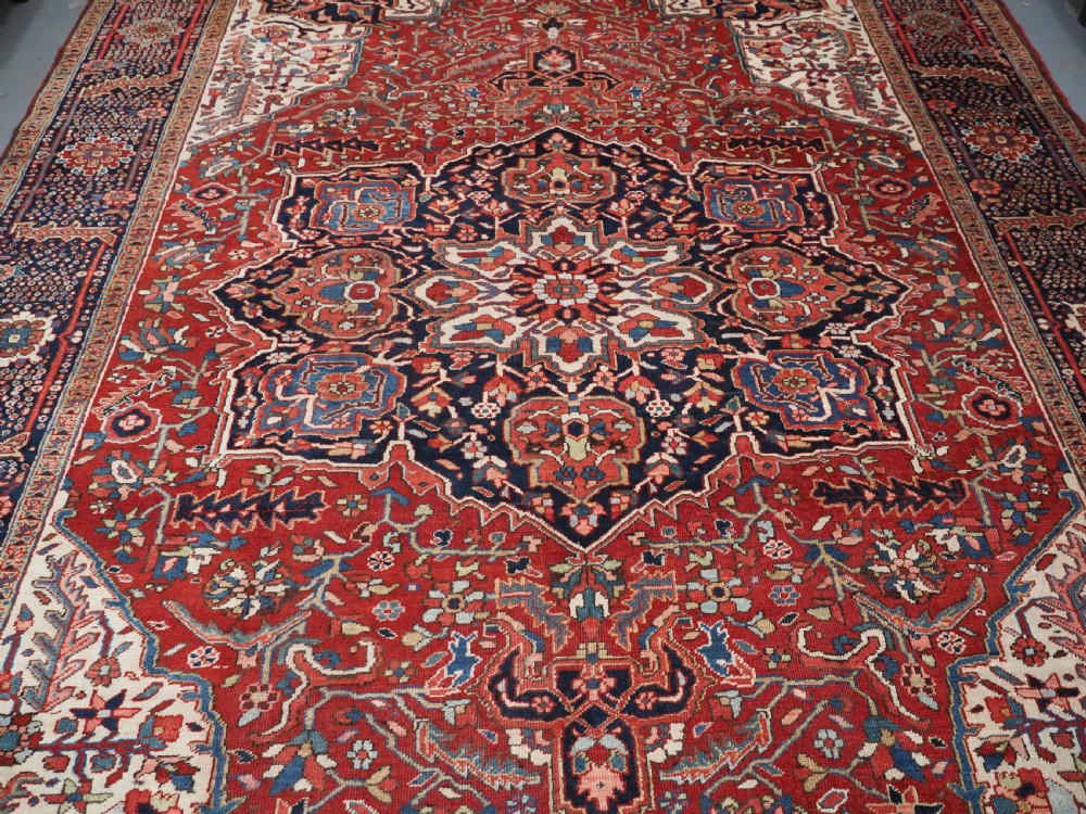An antique hand-knotted Persian Heriz rug featuring a large geometric central medallion in navy blue, set upon a red field with off-white corner spandrels and a wide decorative border. View VI