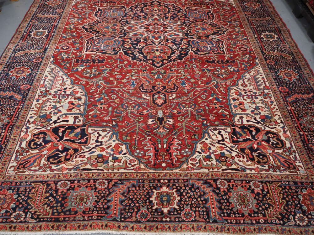 An antique hand-knotted Persian Heriz rug featuring a large geometric central medallion in navy blue, set upon a red field with off-white corner spandrels and a wide decorative border. View VII
