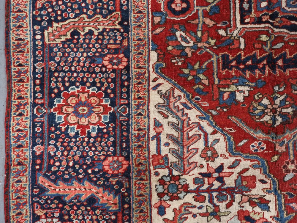 An antique hand-knotted Persian Heriz rug featuring a large geometric central medallion in navy blue, set upon a red field with off-white corner spandrels and a wide decorative border. View IX