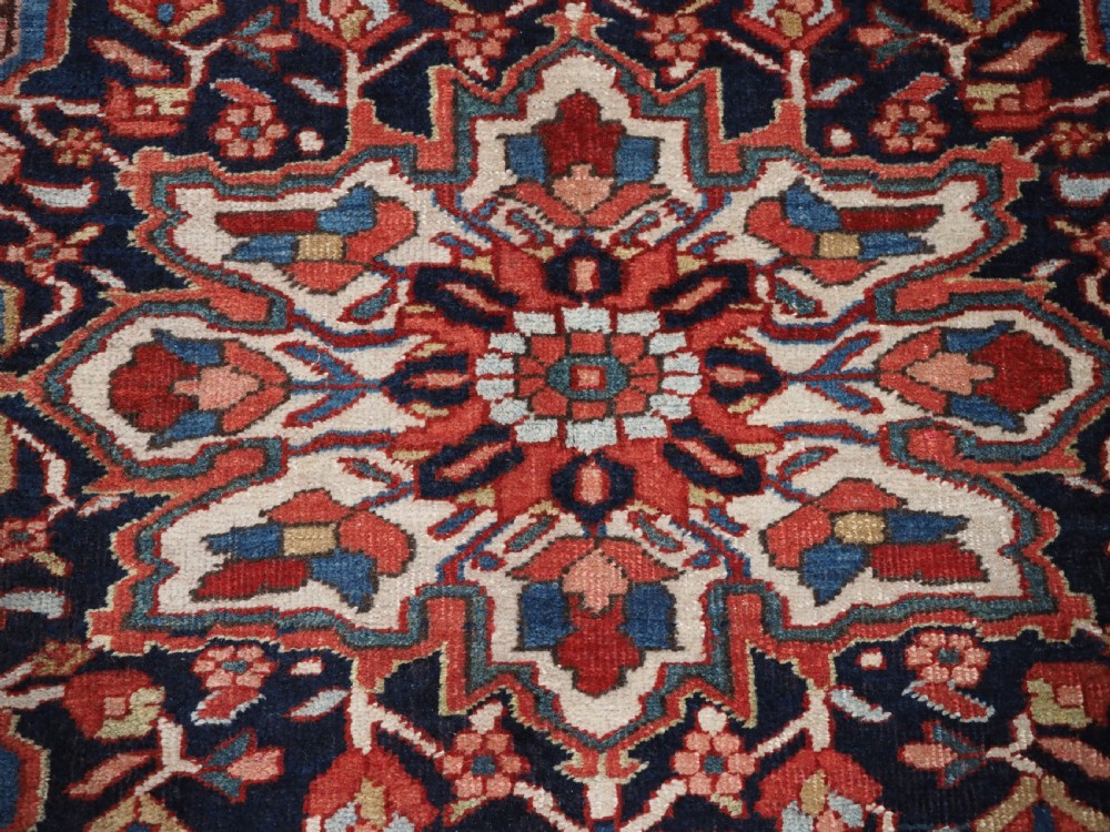 An antique hand-knotted Persian Heriz rug featuring a large geometric central medallion in navy blue, set upon a red field with off-white corner spandrels and a wide decorative border. View XVII