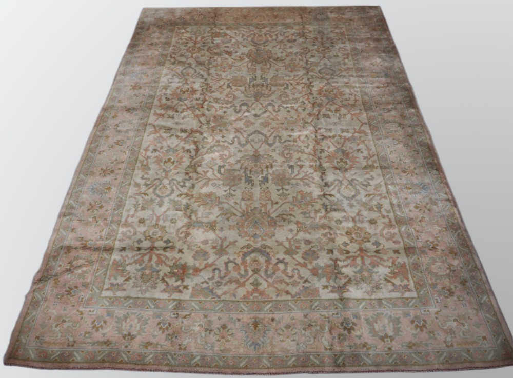 An antique Turkish Oushak wool carpet featuring a soft pink and beige ground decorated with pastel floral palmette patterns and multi-layered borders.
