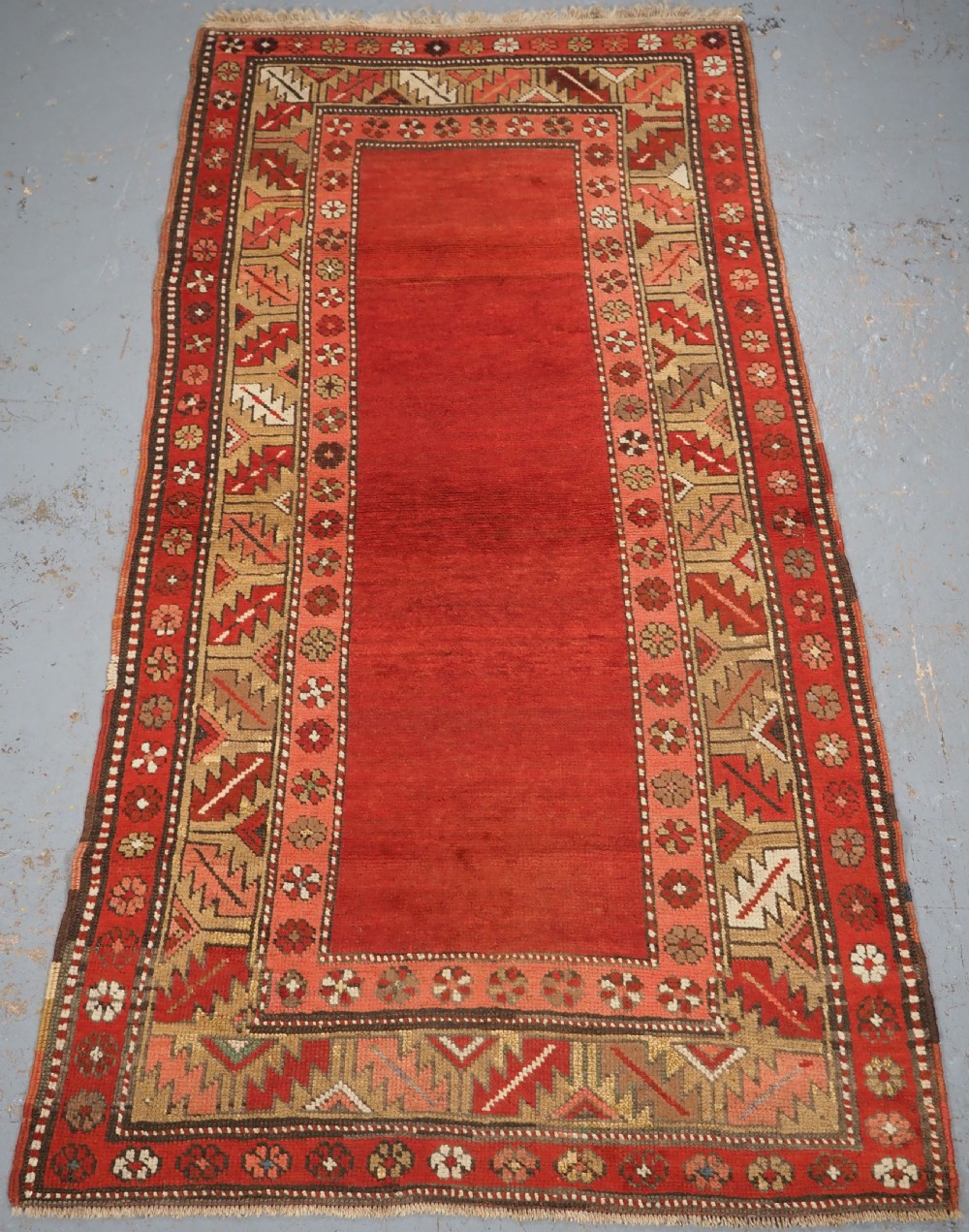 An antique hand-knotted wool rug featuring an unadorned terracotta red field framed by complex geometric and floral patterned borders.