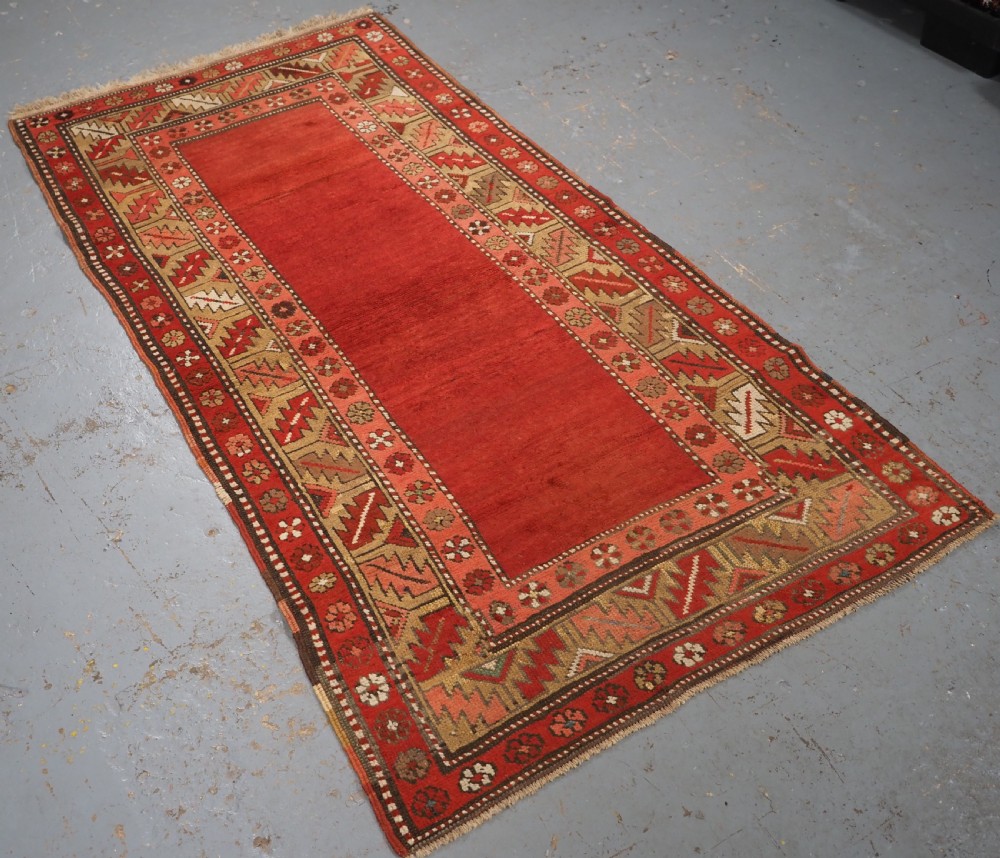 An antique hand-knotted wool rug featuring an unadorned terracotta red field framed by complex geometric and floral patterned borders. View II