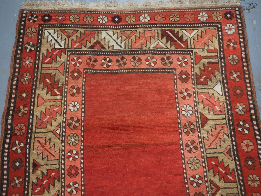 An antique hand-knotted wool rug featuring an unadorned terracotta red field framed by complex geometric and floral patterned borders. View III