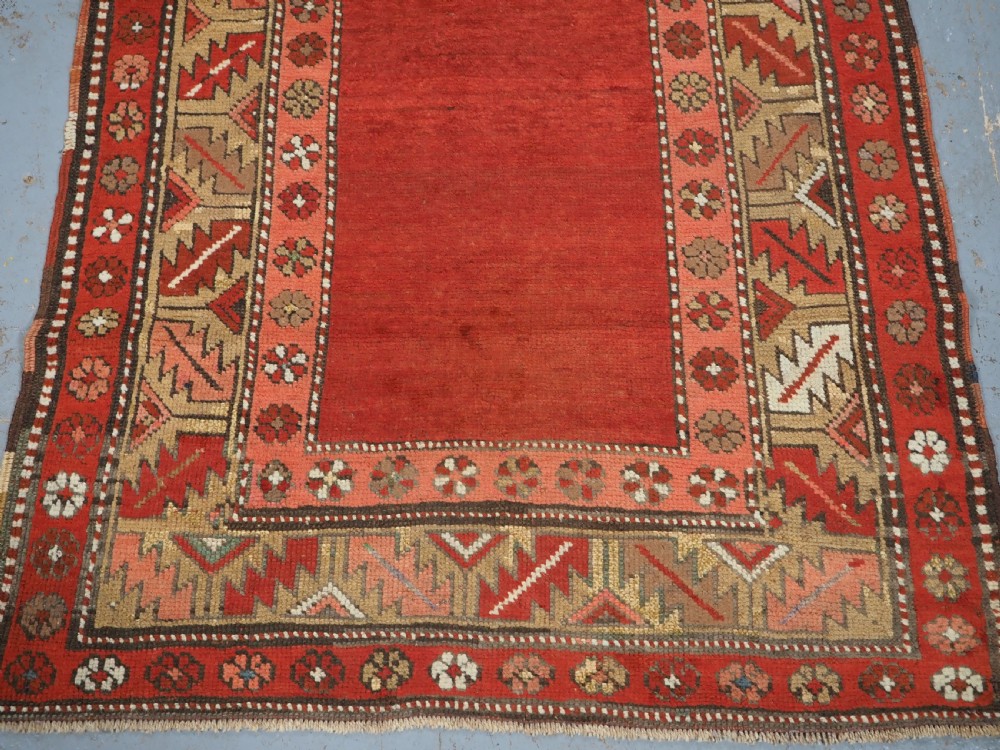 An antique hand-knotted wool rug featuring an unadorned terracotta red field framed by complex geometric and floral patterned borders. View IV