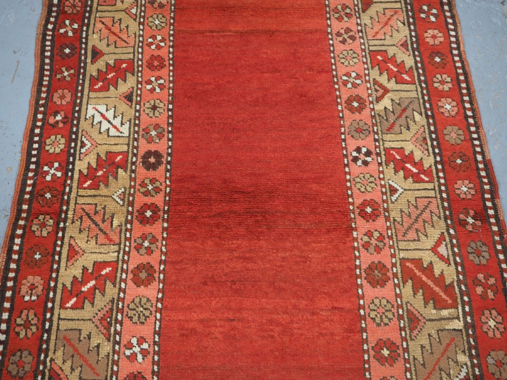 An antique hand-knotted wool rug featuring an unadorned terracotta red field framed by complex geometric and floral patterned borders. View V