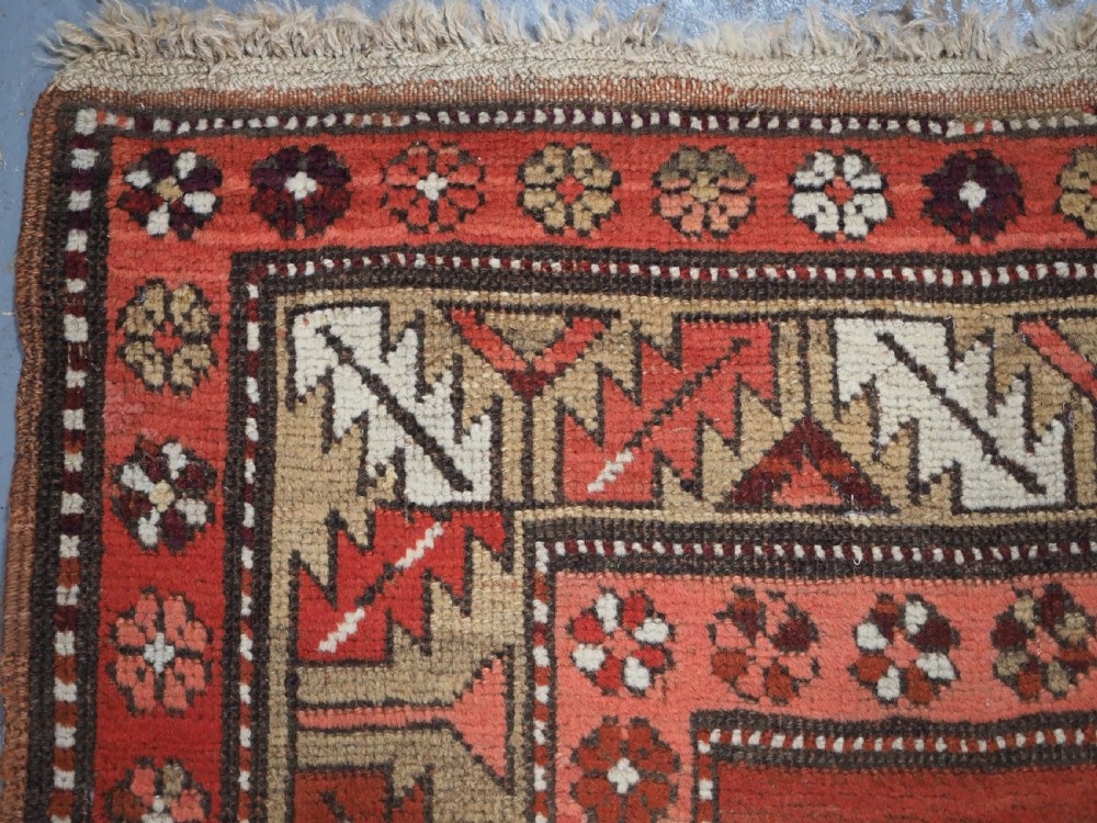 An antique hand-knotted wool rug featuring an unadorned terracotta red field framed by complex geometric and floral patterned borders. View VI
