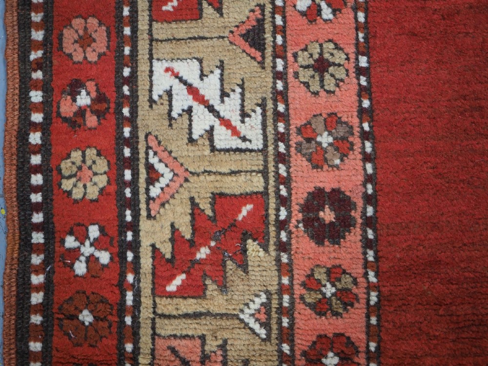 An antique hand-knotted wool rug featuring an unadorned terracotta red field framed by complex geometric and floral patterned borders. View VII