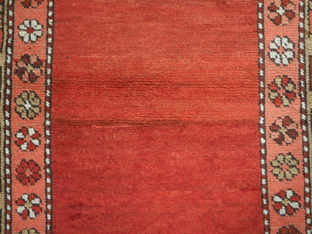 An antique hand-knotted wool rug featuring an unadorned terracotta red field framed by complex geometric and floral patterned borders. View IX
