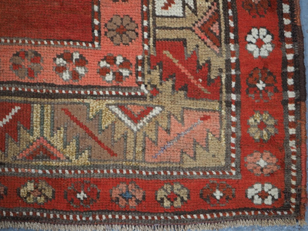 An antique hand-knotted wool rug featuring an unadorned terracotta red field framed by complex geometric and floral patterned borders. View X