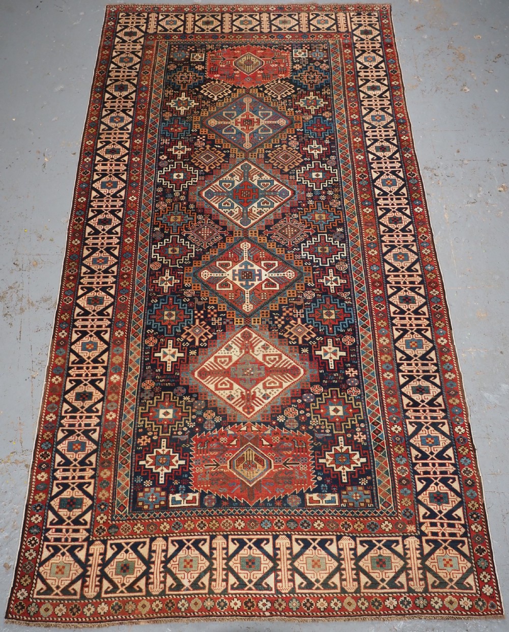 An antique late 19th-century Caucasian Shirvan hand-knotted wool rug featuring geometric diamond medallions, multi-coloured tribal motifs, and a distinct Kufic border.