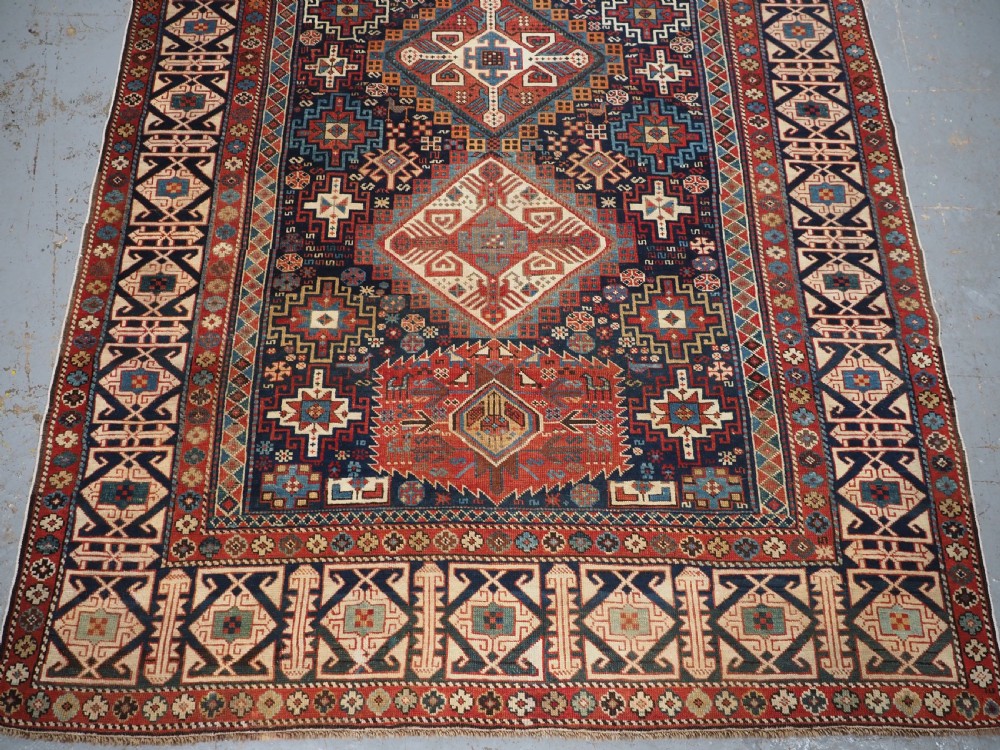 An antique late 19th-century Caucasian Shirvan hand-knotted wool rug featuring geometric diamond medallions, multi-coloured tribal motifs, and a distinct Kufic border. View V