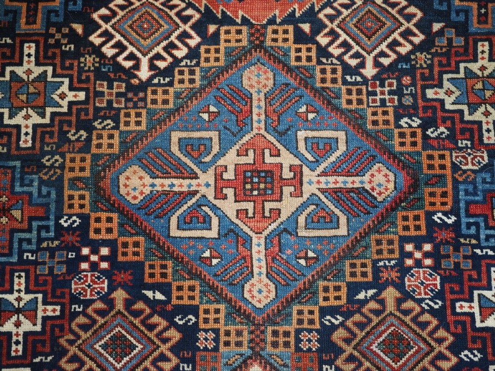 An antique late 19th-century Caucasian Shirvan hand-knotted wool rug featuring geometric diamond medallions, multi-coloured tribal motifs, and a distinct Kufic border. View X