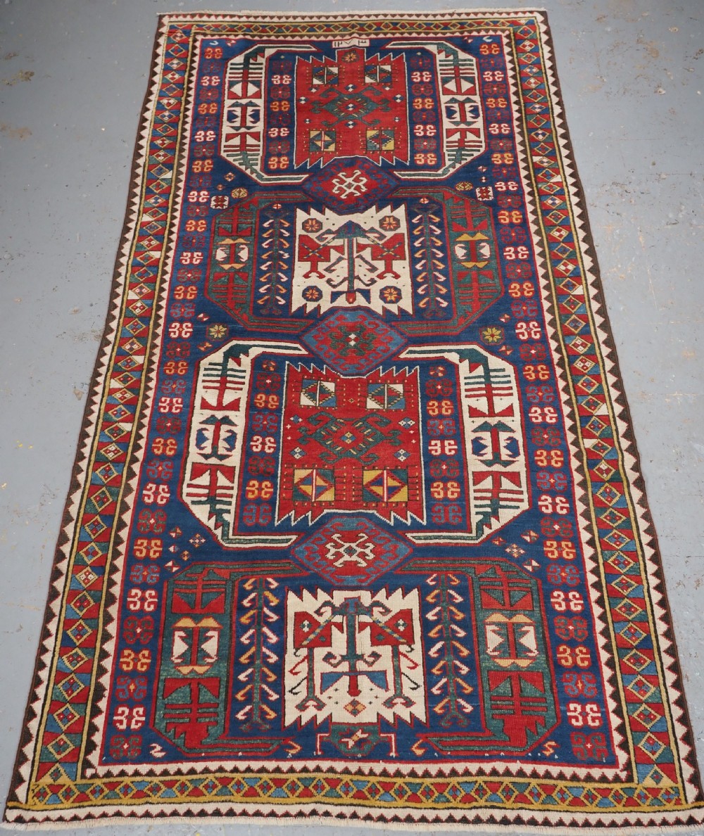 An antique hand-knotted Caucasian Kasim-Usag wool rug featuring geometric red and ivory medallions set on a dark navy blue field with multi-coloured geometric borders.