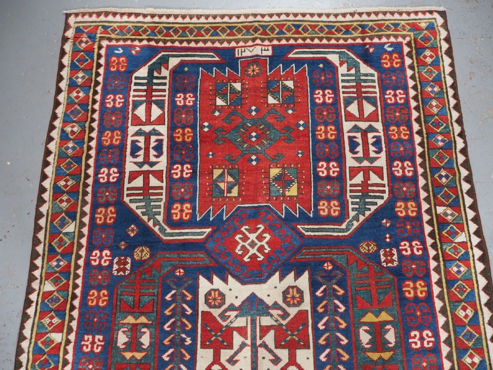 An antique hand-knotted Caucasian Kasim-Usag wool rug featuring geometric red and ivory medallions set on a dark navy blue field with multi-coloured geometric borders. View III
