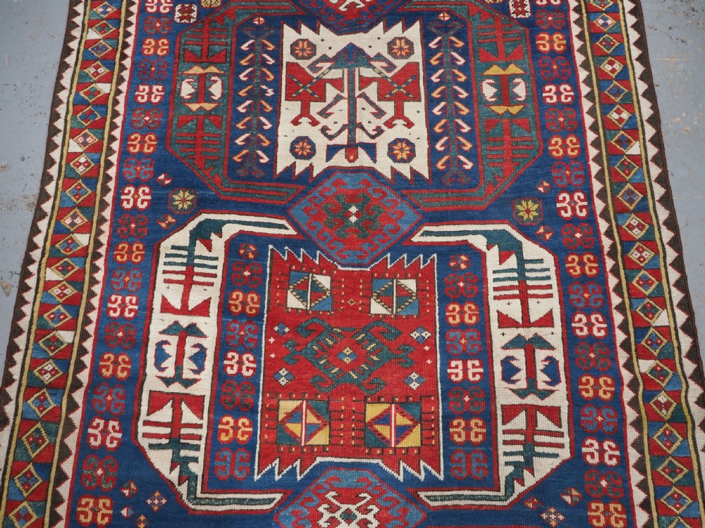 An antique hand-knotted Caucasian Kasim-Usag wool rug featuring geometric red and ivory medallions set on a dark navy blue field with multi-coloured geometric borders. View IV