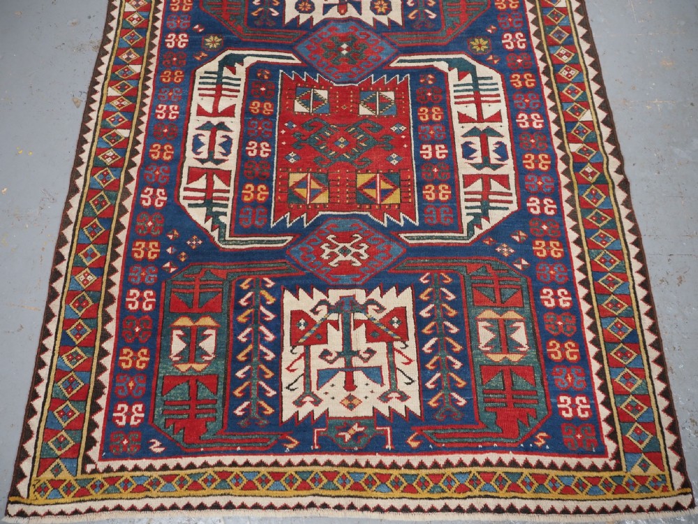 An antique hand-knotted Caucasian Kasim-Usag wool rug featuring geometric red and ivory medallions set on a dark navy blue field with multi-coloured geometric borders. View V