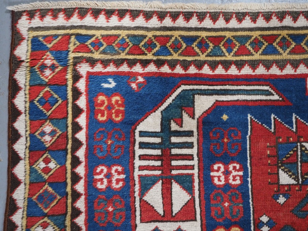 An antique hand-knotted Caucasian Kasim-Usag wool rug featuring geometric red and ivory medallions set on a dark navy blue field with multi-coloured geometric borders. View VI