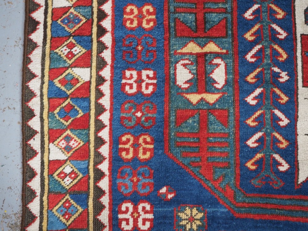 An antique hand-knotted Caucasian Kasim-Usag wool rug featuring geometric red and ivory medallions set on a dark navy blue field with multi-coloured geometric borders. View VII