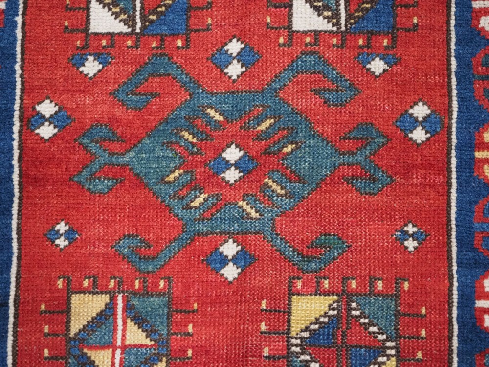 An antique hand-knotted Caucasian Kasim-Usag wool rug featuring geometric red and ivory medallions set on a dark navy blue field with multi-coloured geometric borders. View IX