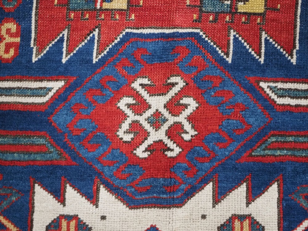 An antique hand-knotted Caucasian Kasim-Usag wool rug featuring geometric red and ivory medallions set on a dark navy blue field with multi-coloured geometric borders. View X