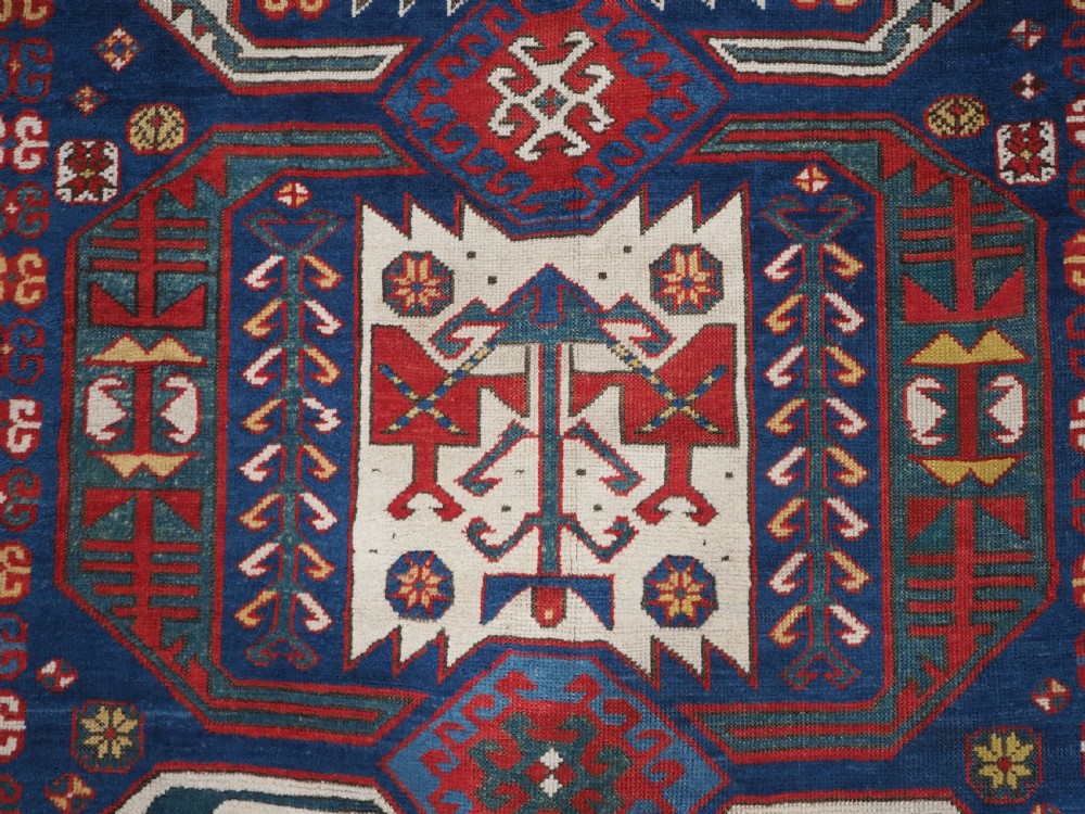 An antique hand-knotted Caucasian Kasim-Usag wool rug featuring geometric red and ivory medallions set on a dark navy blue field with multi-coloured geometric borders. View XI