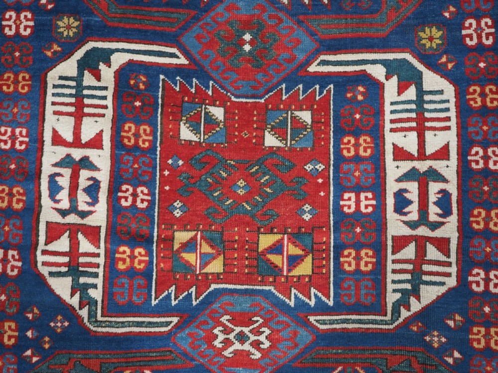 An antique hand-knotted Caucasian Kasim-Usag wool rug featuring geometric red and ivory medallions set on a dark navy blue field with multi-coloured geometric borders. View XII