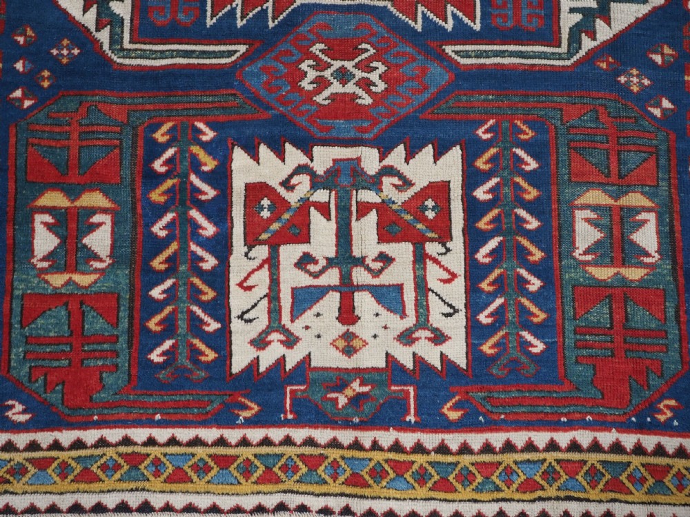 An antique hand-knotted Caucasian Kasim-Usag wool rug featuring geometric red and ivory medallions set on a dark navy blue field with multi-coloured geometric borders. View XIII