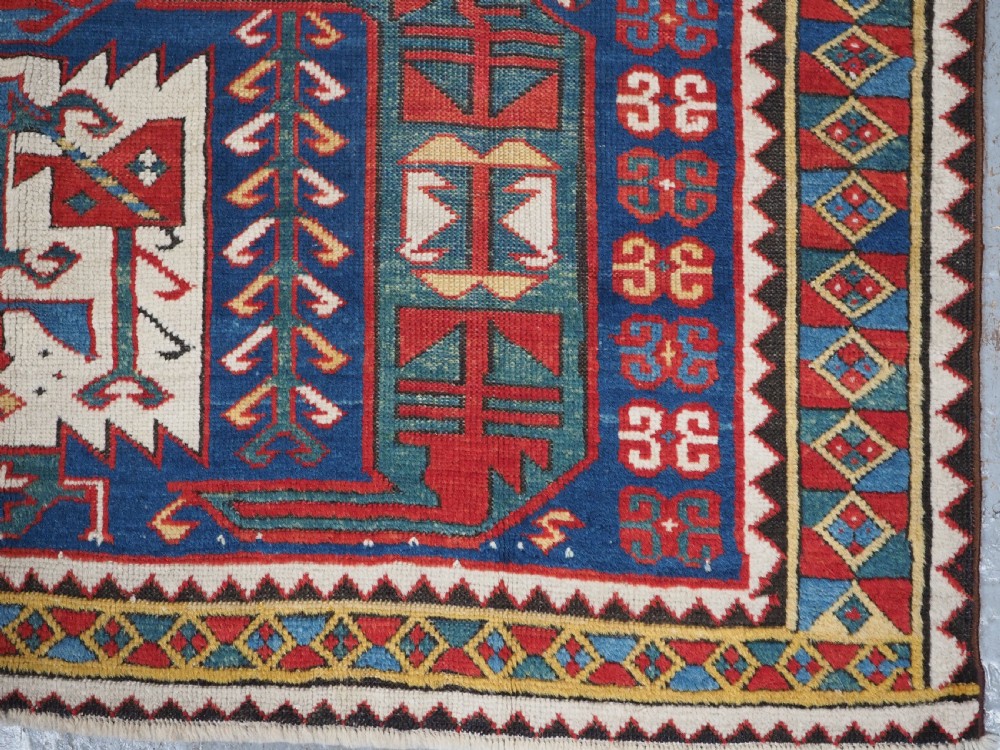 An antique hand-knotted Caucasian Kasim-Usag wool rug featuring geometric red and ivory medallions set on a dark navy blue field with multi-coloured geometric borders. View XIV