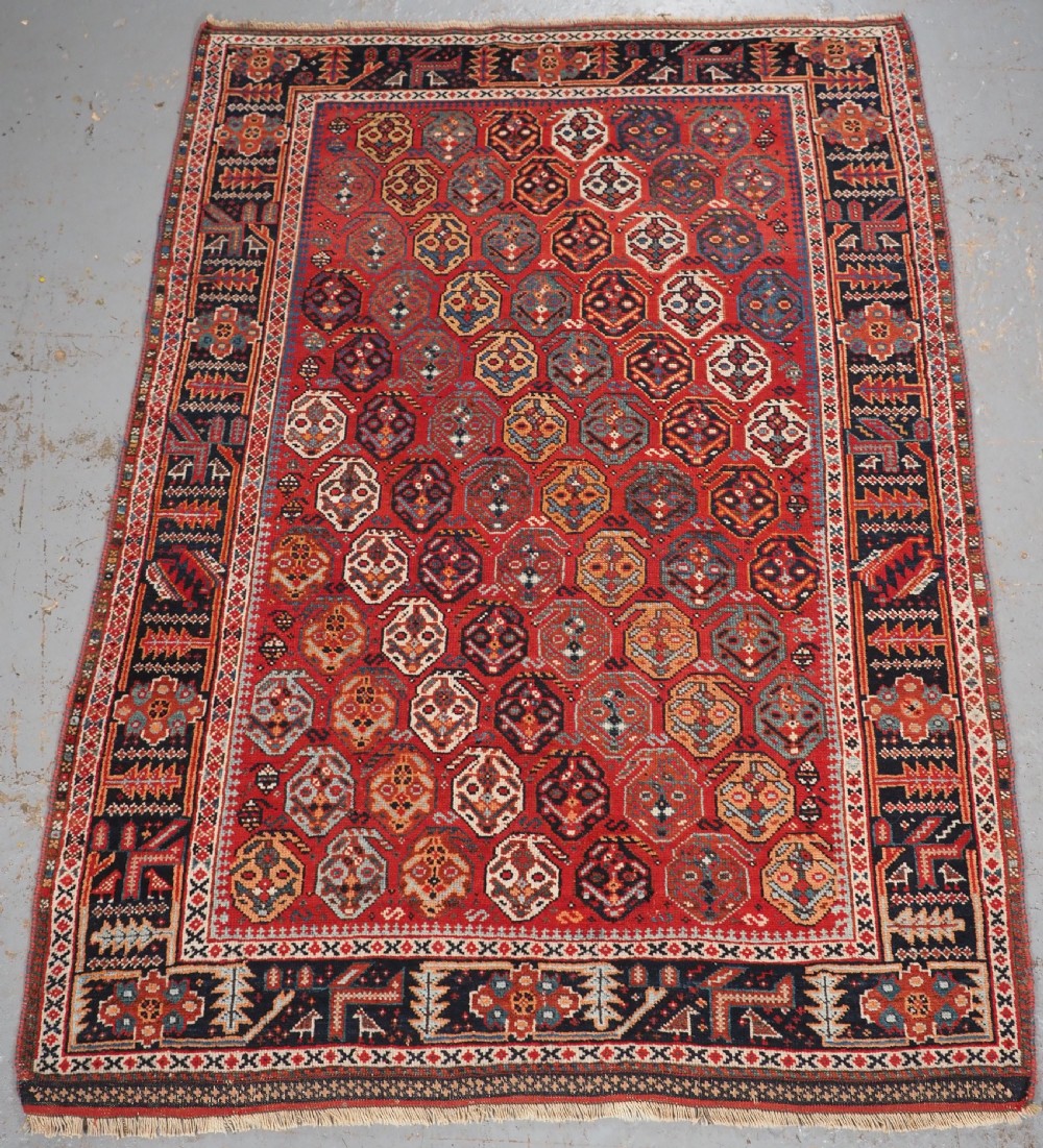 An antique hand-knotted wool rug featuring a deep red field filled with repeating geometric boteh patterns framed by dark blue geometric borders.