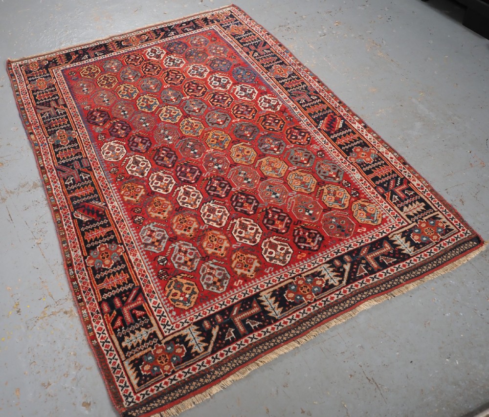 An antique hand-knotted wool rug featuring a deep red field filled with repeating geometric boteh patterns framed by dark blue geometric borders. View II