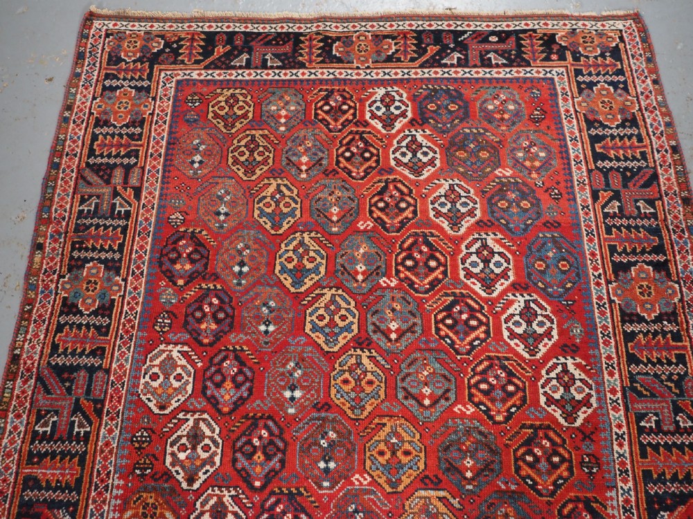An antique hand-knotted wool rug featuring a deep red field filled with repeating geometric boteh patterns framed by dark blue geometric borders. View III
