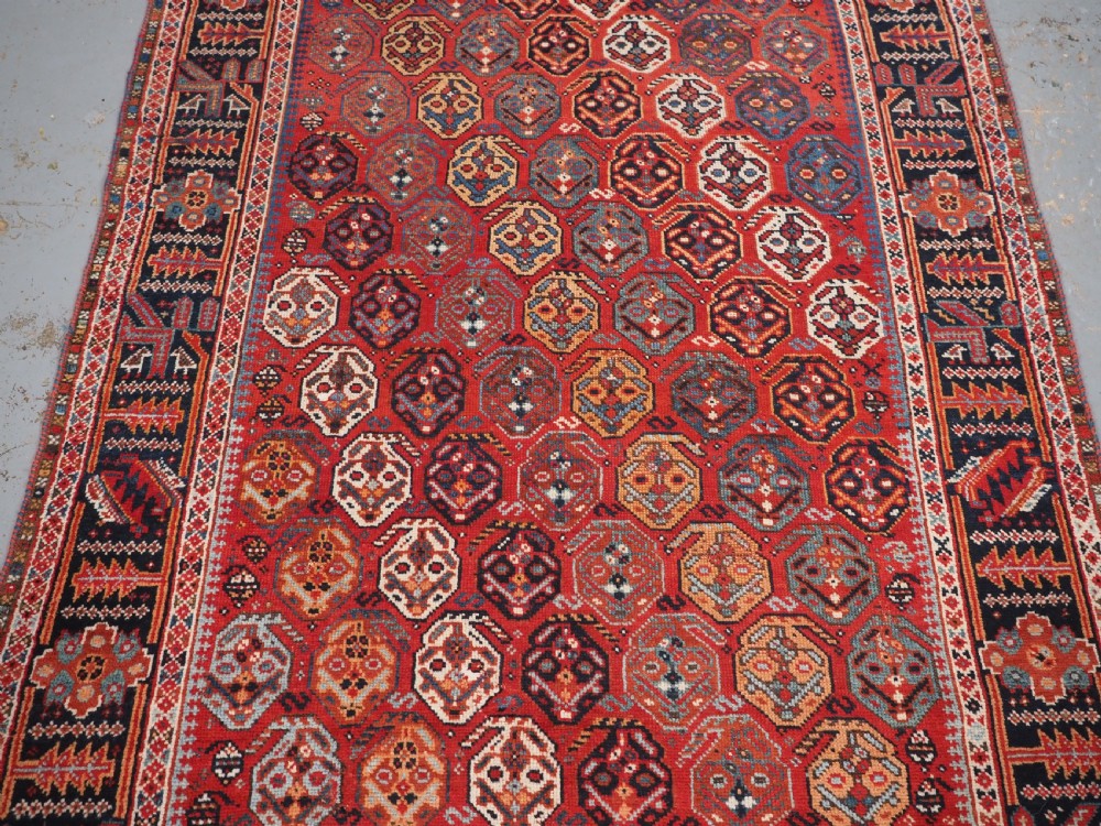 An antique hand-knotted wool rug featuring a deep red field filled with repeating geometric boteh patterns framed by dark blue geometric borders. View IV