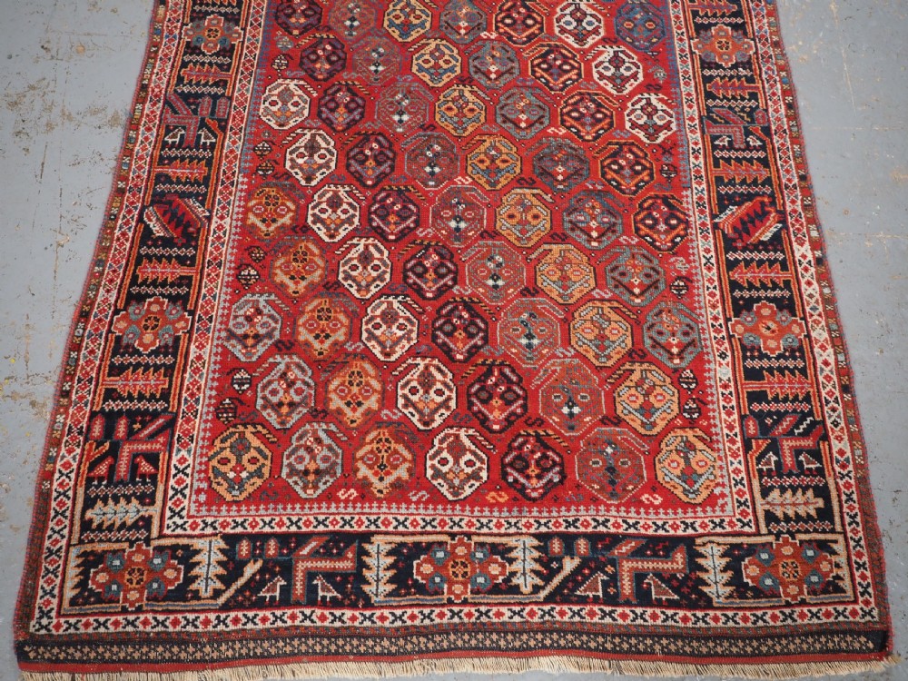 An antique hand-knotted wool rug featuring a deep red field filled with repeating geometric boteh patterns framed by dark blue geometric borders. View V