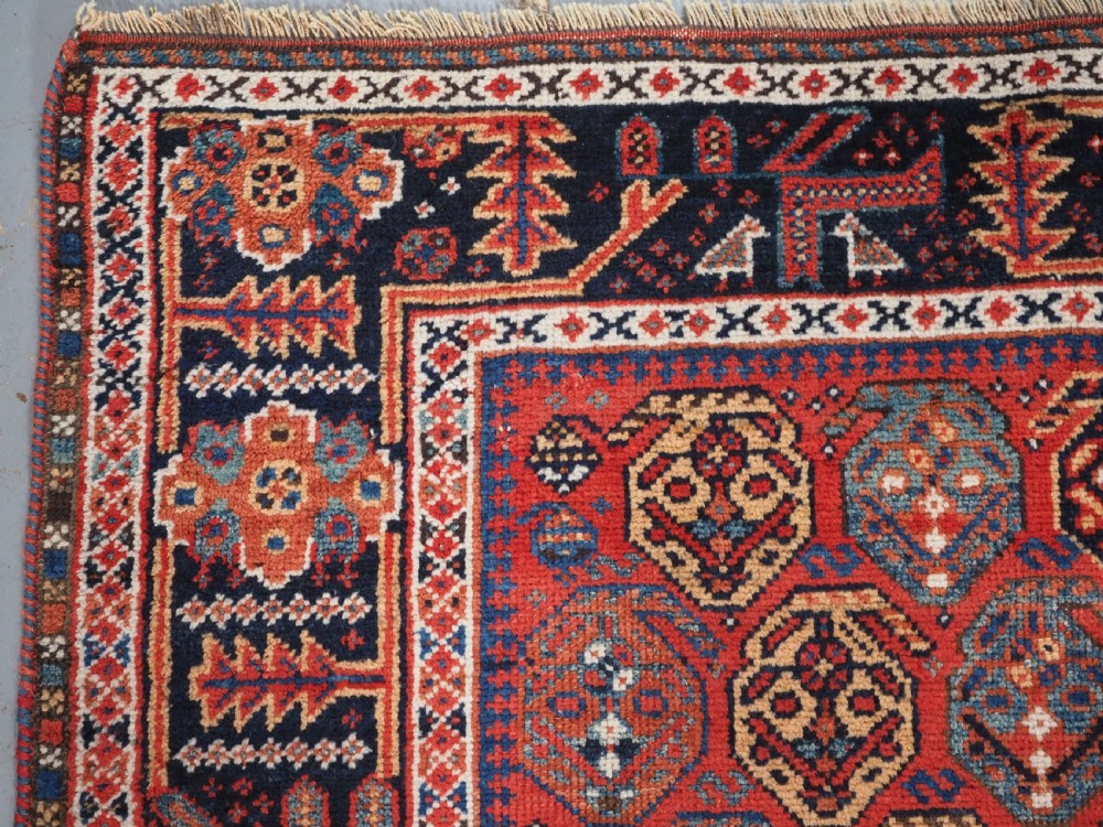 An antique hand-knotted wool rug featuring a deep red field filled with repeating geometric boteh patterns framed by dark blue geometric borders. View VI