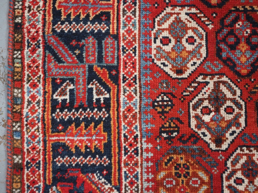 An antique hand-knotted wool rug featuring a deep red field filled with repeating geometric boteh patterns framed by dark blue geometric borders. View VII