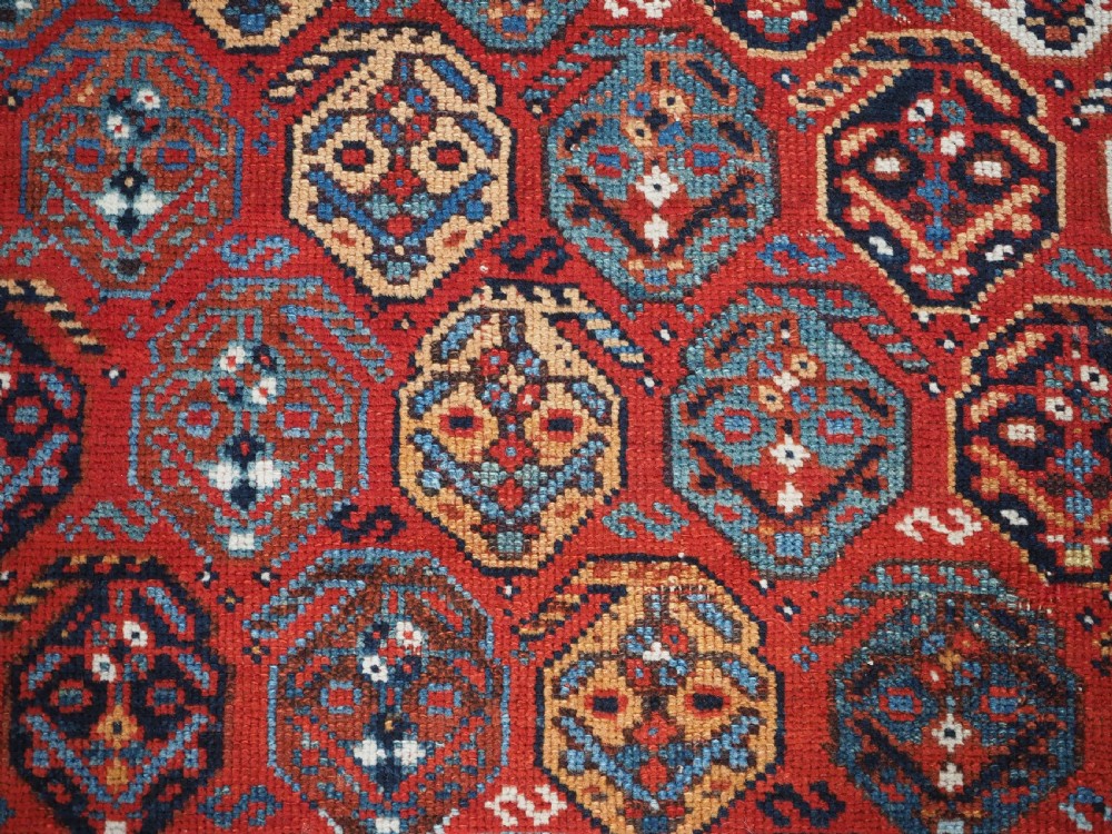 An antique hand-knotted wool rug featuring a deep red field filled with repeating geometric boteh patterns framed by dark blue geometric borders. View IX