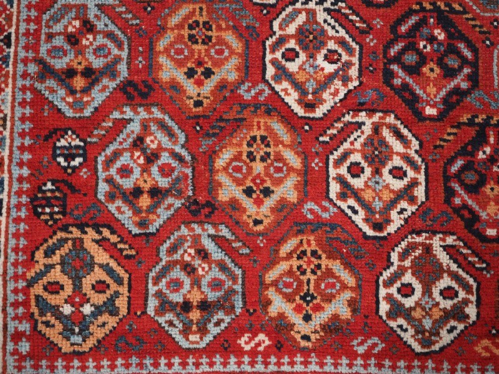 An antique hand-knotted wool rug featuring a deep red field filled with repeating geometric boteh patterns framed by dark blue geometric borders. View X
