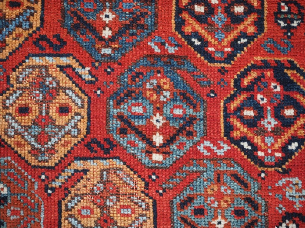 An antique hand-knotted wool rug featuring a deep red field filled with repeating geometric boteh patterns framed by dark blue geometric borders. View XI