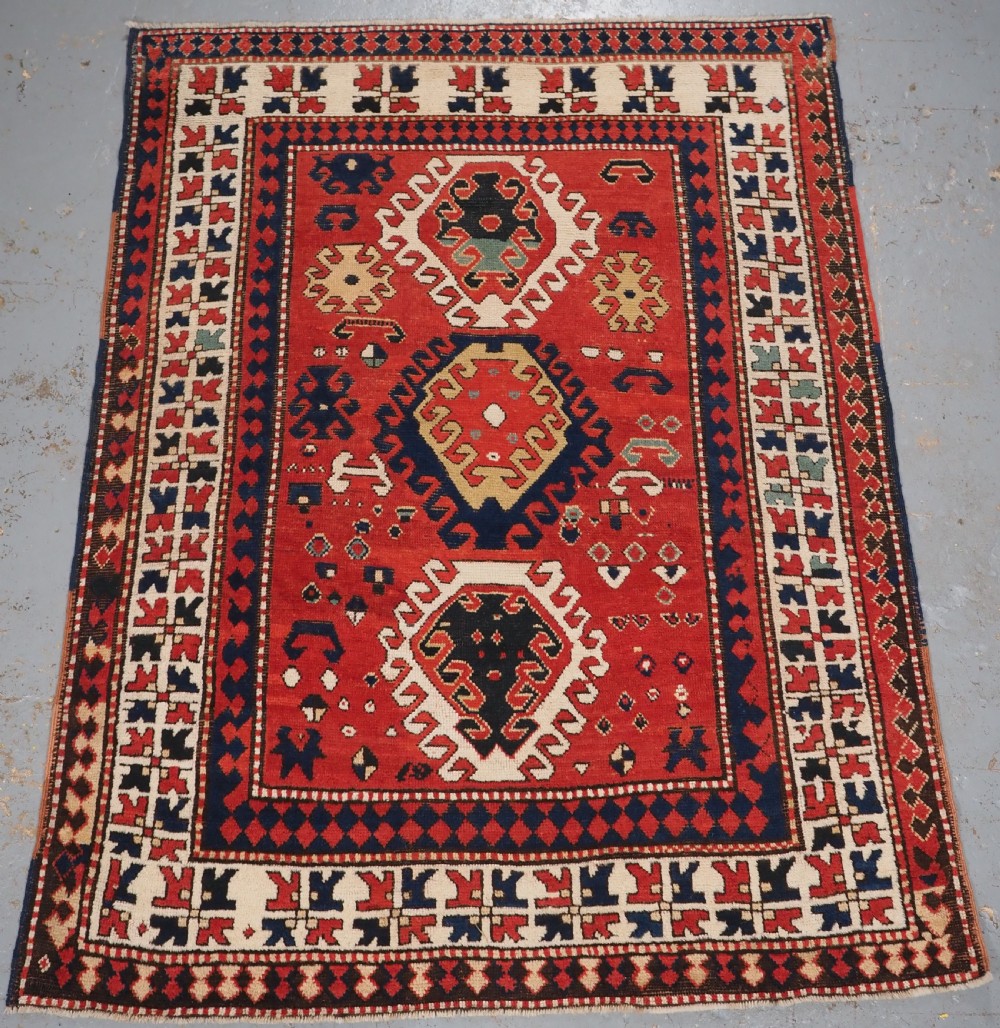 An antique Caucasian Bordjalou Kazak hand-knotted wool rug featuring three stepped geometric medallions on a vibrant red field within patterned borders.