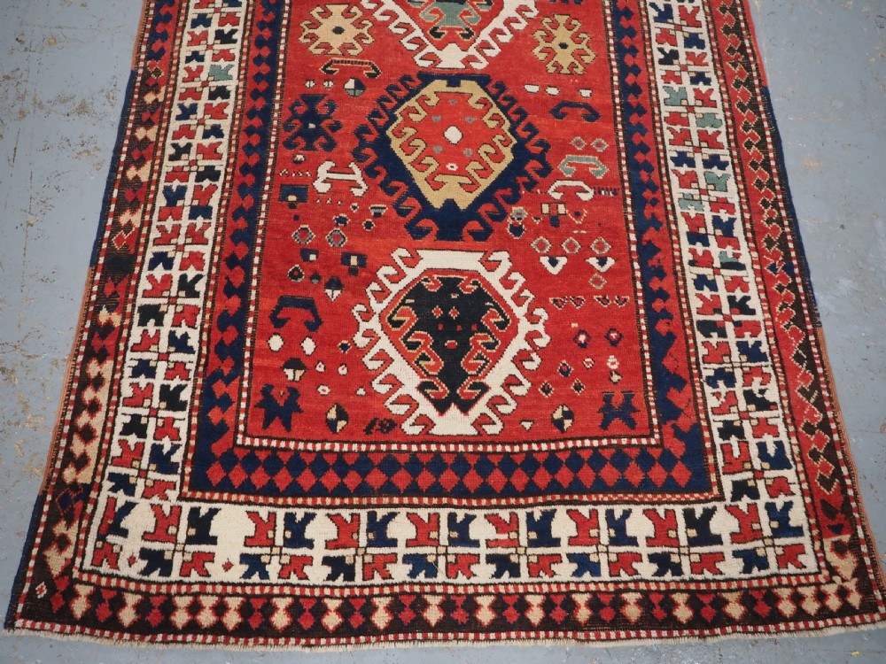 An antique Caucasian Bordjalou Kazak hand-knotted wool rug featuring three stepped geometric medallions on a vibrant red field within patterned borders. View V