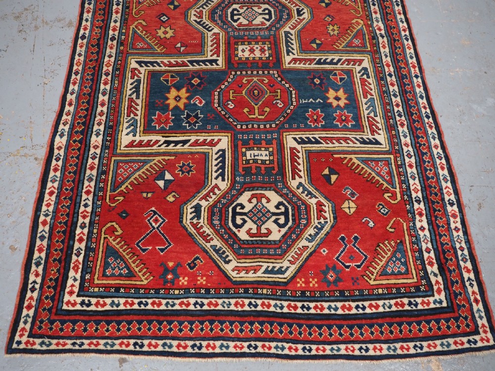 An antique Caucasian Sewan Kazak wool rug featuring a red keyhole medallion set against a navy blue and cream background with geometric borders. View V
