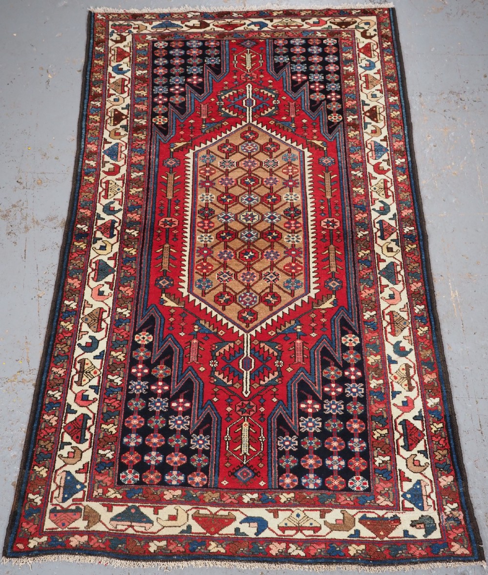 An antique hand-knotted Persian Mazlaghan wool rug featuring a central camel-coloured medallion on a red stepped field with dark blue rosette borders.